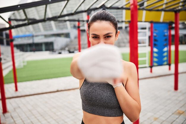 Waist up portrait of athletic young female in boxing bandages demonstrating cross hit to camera