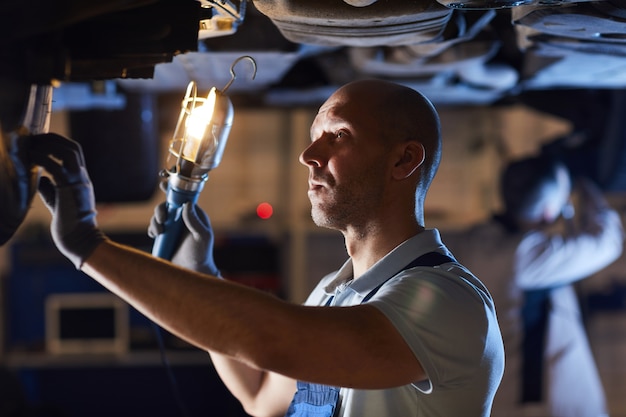 Waist up portrait of bald car mechanic inspecting vehicle while holding flashlight lamp under car lift, copy space