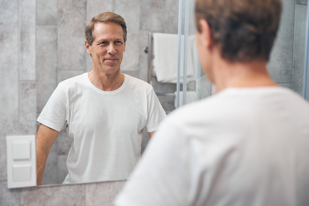 Waist up portrait of an  blond adult man standing in front of the mirror