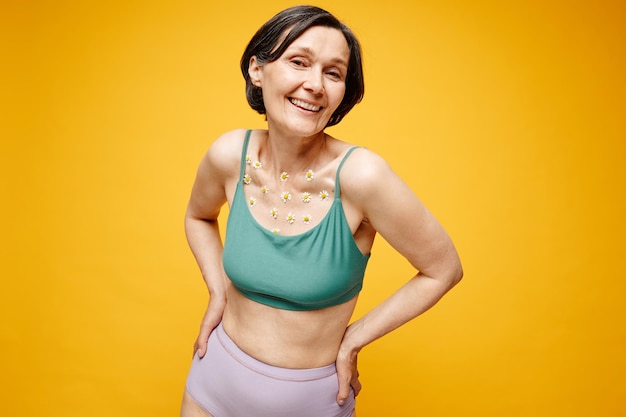 Waist up portrait of carefree mature woman with flowers on skin wearing underwear against vibrant ye