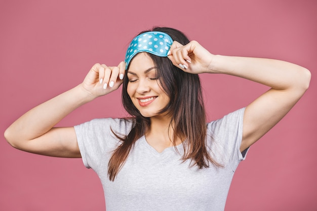 Waist up portrait of cheerful smiling girl in sleeping mask. Attractive funny female in stylish pajama standing and looking away. Isolated on pink background.