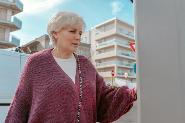 Waist-up portrait of a concentrated gray-haired lady using the self-service payment terminal at the modern gas station