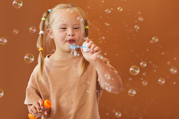 Waist up portrait of cute girl with down syndrome blowing bubbles while posing against brown surface in studio, copy space