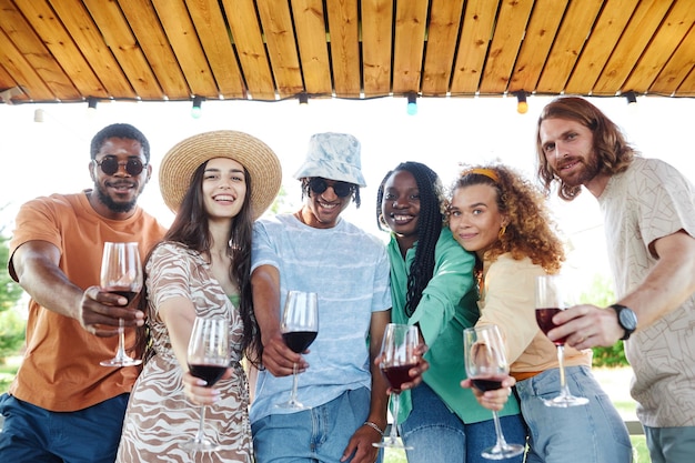 Waist up portrait of diverse group of friends looking at camera and holding wine glasses while enjoy