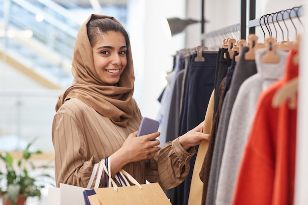 Waist up portrait of elegant young woman  while standing by clothing racks and enjoying shopping in mall