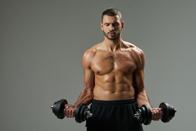 Waist up portrait of focused bearded strong Caucasian sportsman in black sportswear lifting weights in room indoors