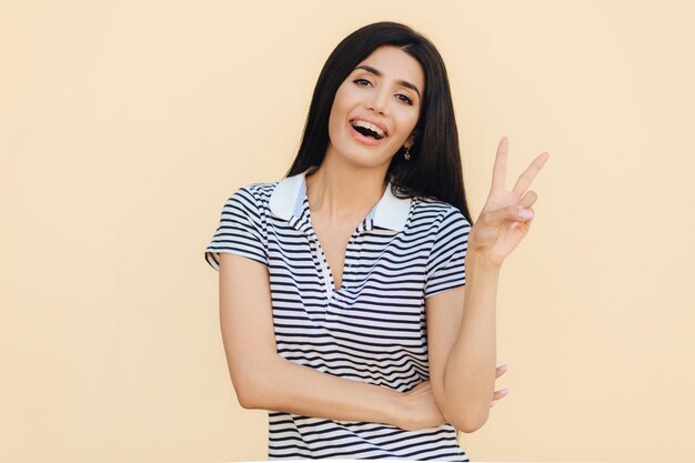 Waist up portrait of happy brunette young female shows peace sign smiles happily at camera wears braces on teeth has friendly look models indoor People body language and happiness concept