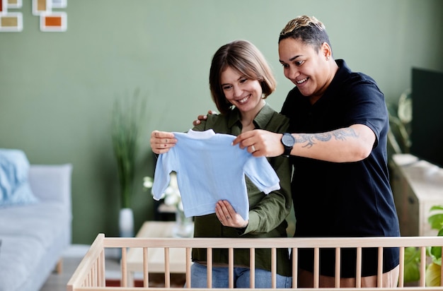 Waist up portrait of happy lesbian couple expecting baby and preparing crib with baby clothes at hom