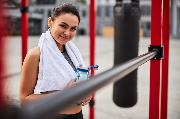 Waist up portrait of happy slim female with towel and bottle of water standing on ring in open air