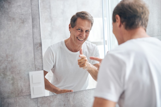 Waist-up portrait of a high-spirited short-haired mature man pointing his finger at the mirror