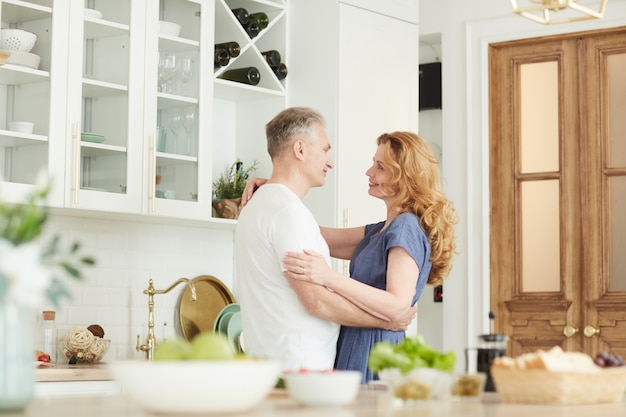 Waist up portrait of loving mature couple embracing and looking at each other in white kitchen interior at home, copy space