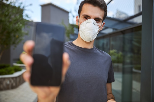 Waist-up portrait of a male in a protective mask holding his cellphone in front of the camera