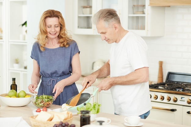 Waist up portrait of modern mature couple enjoying cooking together at home in white kitchen, interior copy space