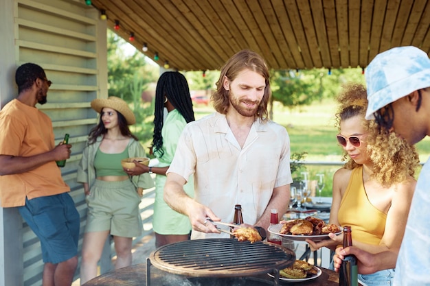 Waist up portrait of smiling man grilling meat outdoors during summer barbeque party with friends