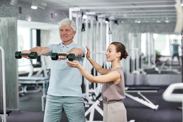 Waist up portrait of smiling senior man doing physical exercises in rehabilitation clinic with femal