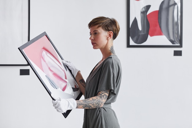 Waist up portrait of tattooed creative woman holding painting while planning art gallery exhibition, 