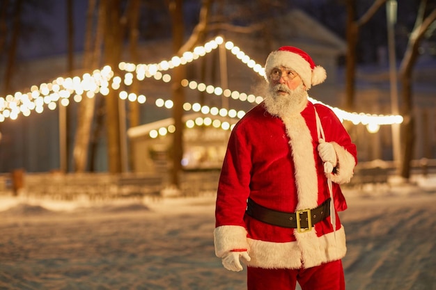 Waist up portrait of traditional santa claus carrying bag with presents at night with fairy lights i