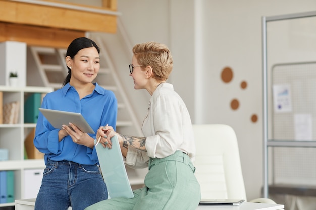 Waist up portrait of two modern businesswomen chatting cheerfully while enjoying work in office, copy space