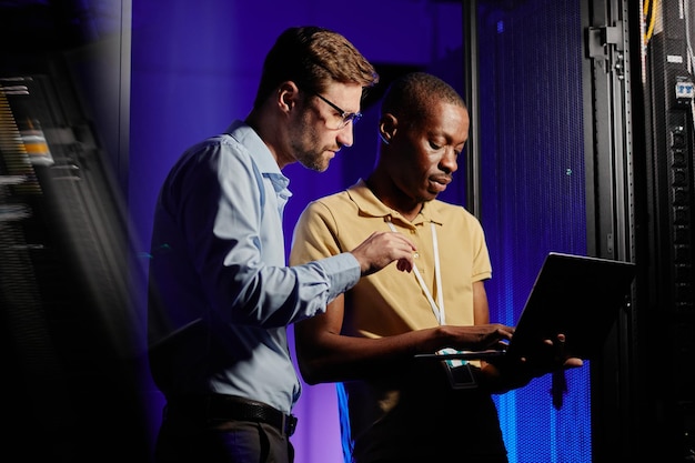 Waist up portrait of two network engineers using computer in dark server room lit by neon light copy