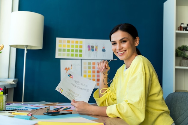 Waist up portrait view of the brunette female designer smiling to the camera while holding her sketches of future clothes collection during sitting at her workplace