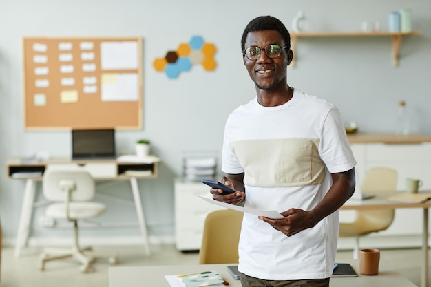 Waist up portrait of young black man wearing casual clothes in it office and holding smartphone faci