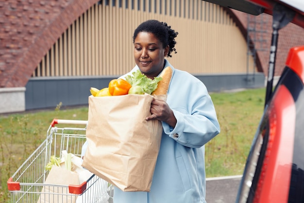 Waist up portrait of young black woman putting grocery bag in car trunk at supermarket parking lot