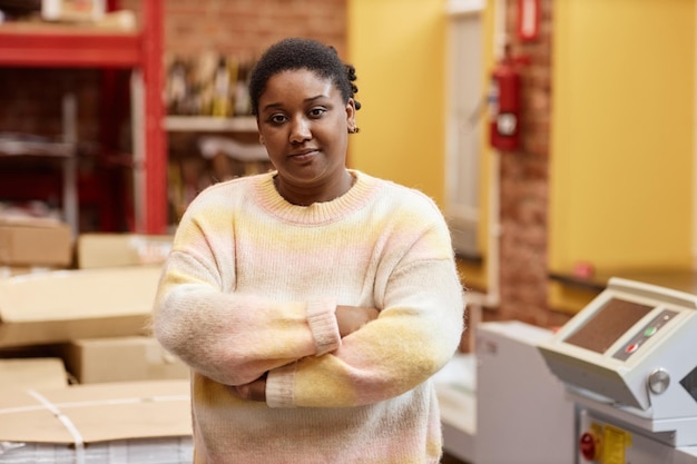 Waist up portrait of young black woman working in industrial printing shop and looking at camera whi