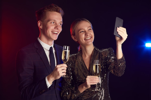 Waist up portrait of young couple taking selfie photo via smartphone while enjoying party at prom night, copy space