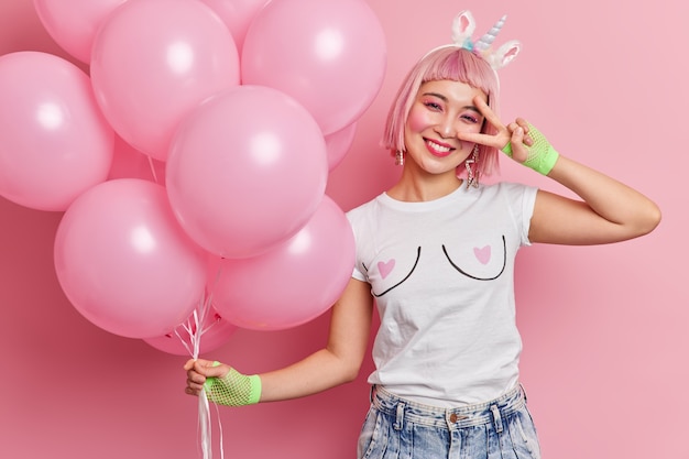 Waist up shot of Asian pink haired woman makes peace gesture over eye smiles pleasantly dressed in casual t shirt and jeans has fun on party. People joy entertainment and celebration concept