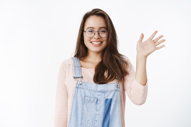 Waist-up shot of cute female newbie waving new coworker hopefully, making friends smiling friendly and happy as raising palm in hello or hi gesture, greeting everyone pleased and joyful.