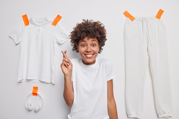 Waist up shot of happy beautiful woman with dark curly hair keeps fingers crossed makes wish dressed in casual t shirt poses against white background with plastered clothes Good luck concept