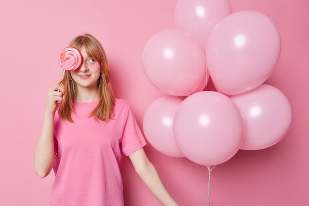 Waist up shot redhead girl dressed in casual t shirt covers eye with lollipop holds bunch of inflated balloons celebrates special occasion isolated over pink background Ginger female with candy