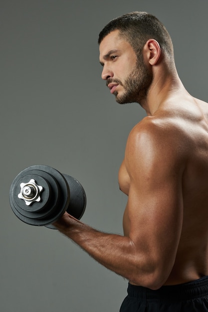 Waist up side view portrait of attractive Caucasian sportsman in black sportswear holding dumbbell in hand while looking away in the indoors