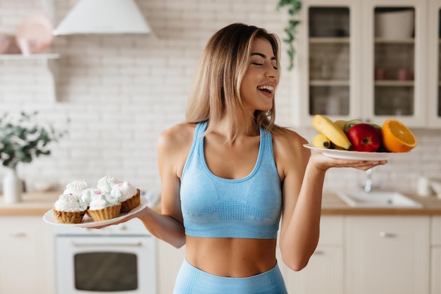 Waistup of energetic young lady posing in her kitchen and holding plates with fruit and desserts