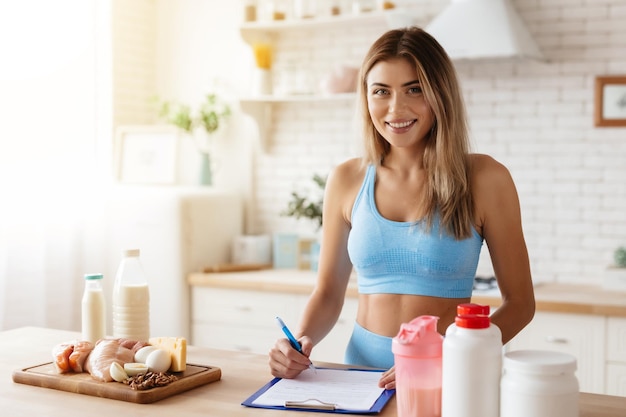 Waistup photo of smiley young woman taking notes while standing in kitchen near food supplement containers and some products