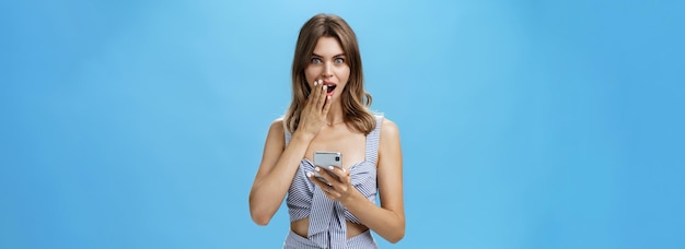 Waistup shot of surprised and excited attractive woman holding smartphone covering opened mouth with