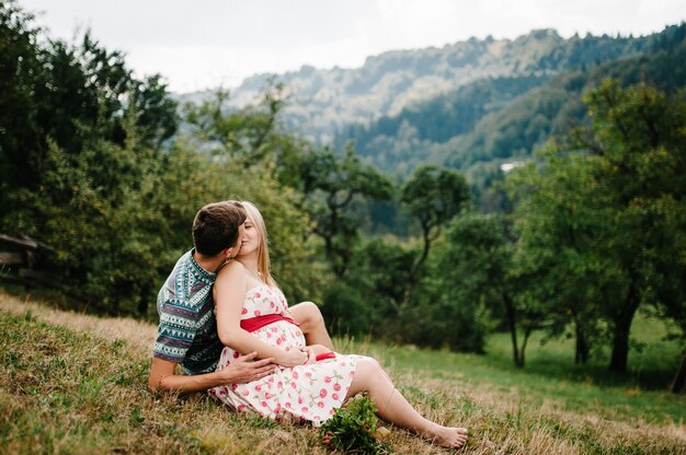 Wait baby. happy family. pregnant woman with beloved husband sitting on grass, kissing. hand embraces round belly. sincere tender moment. background, nature, park, tree, mountain, forest, nine month