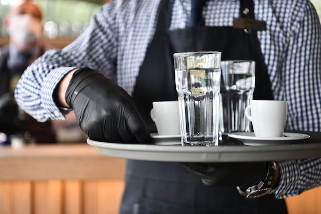 Photo waiter in a medical protective mask serves  the coffee in restaurant durin coronavirus pandemic representing new normal concept