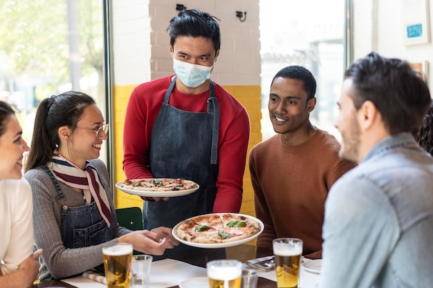 Waiter in protective face mask serving delicious pizzas margherita to friends in cozy pizzeria restaurant Multiethnic friends having fun together in the pizzeria eating pizza and drinking blond beer