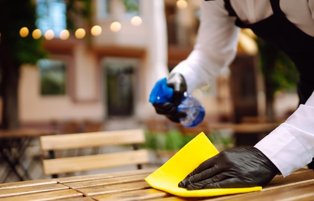 Waiter in protective mask and gloves disinfecting restaurant table for next customer.