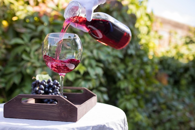 A waiter's hand in a white glove pours a glass of red wine which stands on a table with a white tablecloth outdoors in the sunlight