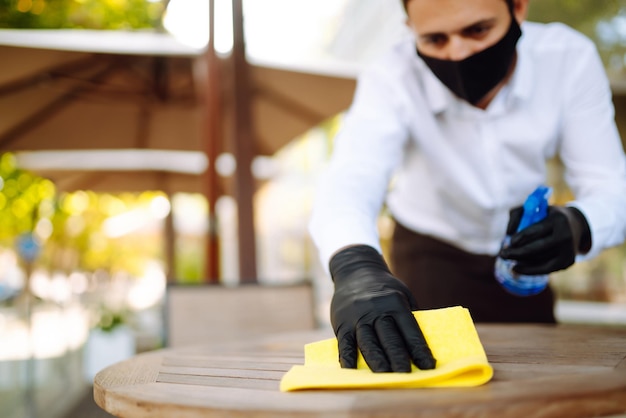 Photo waiter wearing protective face mask and gloves while disinfecting tables at outdoor cafe