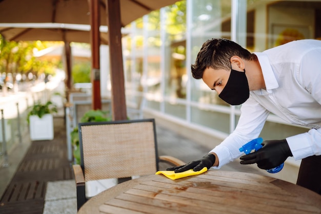 Waiter wearing protective face mask and gloves while disinfecting tables at outdoor cafe