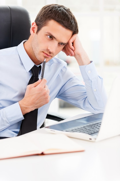 Waiting for inspiration. Thoughtful young man in shirt and tie looking at laptop and touching his chin with pen while sitting at his working place