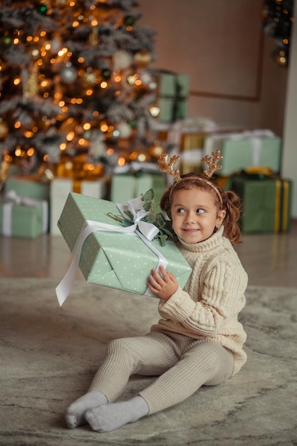 Waiting for the new emotional portrait of a little girl at home in a room near the Christmas tree with a gift in her hands