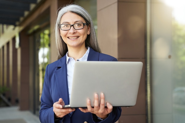 Waiting for a partner. Portrait of beautiful mature business woman wearing classic suit and eyeglasses looking at laptop screen while standing against office building outdoors. Business people at work