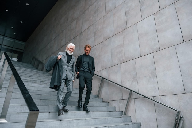 Walking on the stairs Young guy with senior man in elegant clothes is outdoors together Conception of business