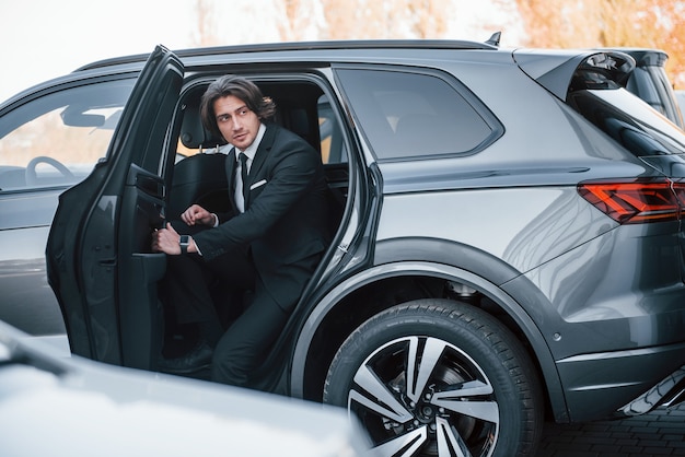 Walks out from car. Young businessman in black suit and tie inside modern automobile.