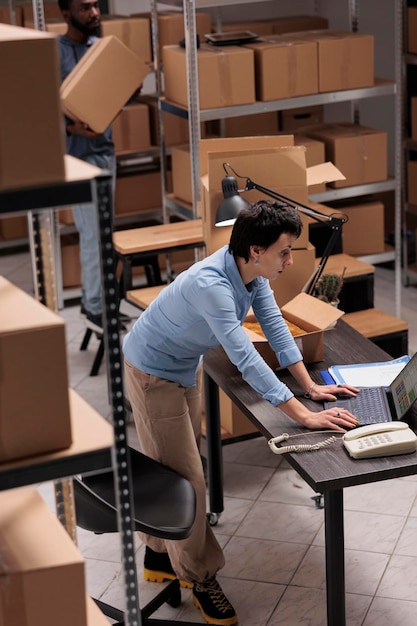 Warehouse manager checking shipping details on laptop computer before start packing customers orders, putting clothes in carton boxes, preparing for delivery. Woman working in delivery department