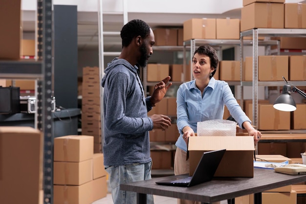 Warehouse team discussing about distribution logistics while putting client goods in carton box after wrapping order in bubble wrap. Industrial employee working with merchandise in storehouse